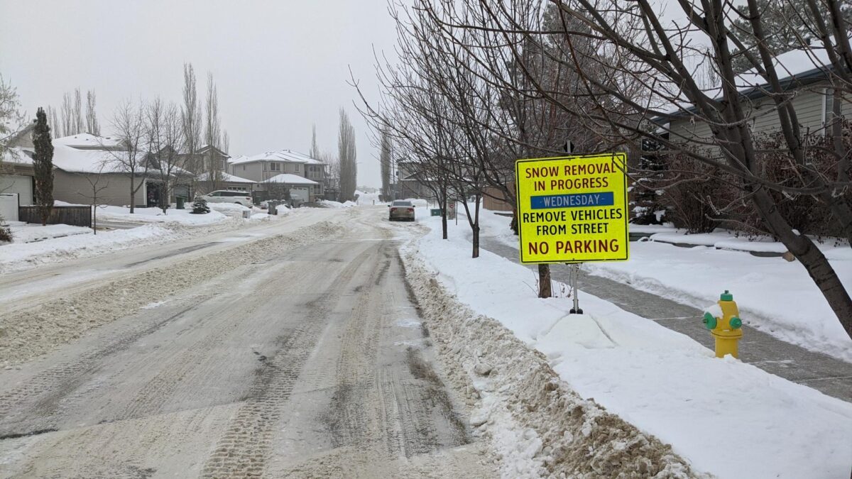 Image of a snowy street in Beaumont that has been partially cleared of slow. The focus is a yellow sign indicating snow removal in progress.