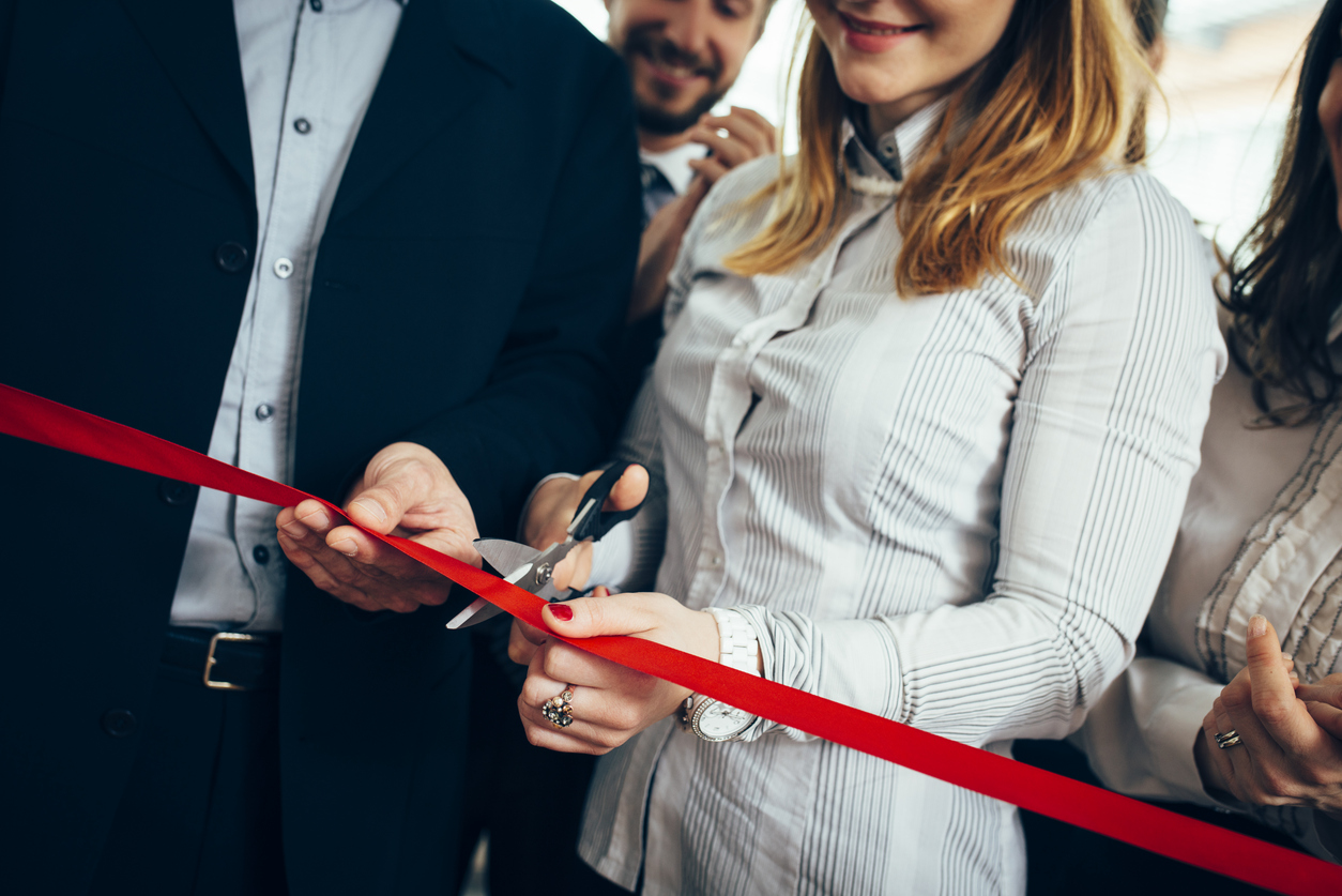 Photo of a ribbon cutting with a red ribbon pulled diagonally across the width of the image and a masculine figure holding the ribbon for a feminine figure to cut it.