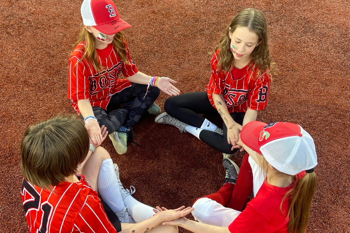 Four girls in baseball uniforms sitting in a circle on the shale surface of the baseball diamond.